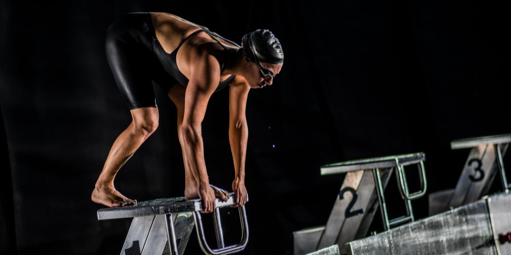 Eine Frau in einem schwarzen Badeanzug steht in Starposition auf einem Startblock
