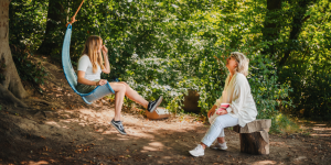 Daniela Brandl sitzt auf einem Holzstamm im Wald. Vor ihr eine Klientin, die auf einen blauen Schaukel sitzt, die an einem Baum hängt.