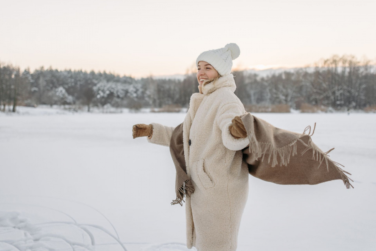 Eine Frau in einem weißen Wollmantel mit beidem Tuch darüber steht in einer Schneelandschaft. Sie hat die Arme ausgebreitet.