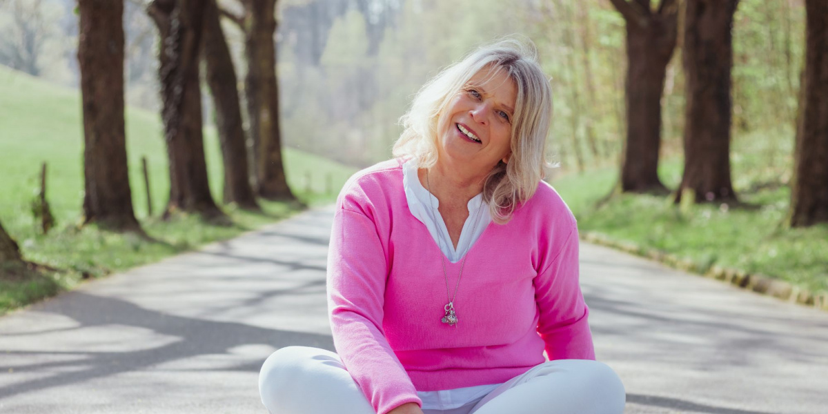 Daniela Brandl, Coach für Frauen und Business, sitzt lachend im Schneidersitz auf einer von Bäumen gesäumten Landstraße. Sie trägt einen pinkfarbenen Pullover, wirkt entspannt, lebensfroh und ganz bei sich – ein Moment voller Leichtigkeit und Naturverbundenheit.