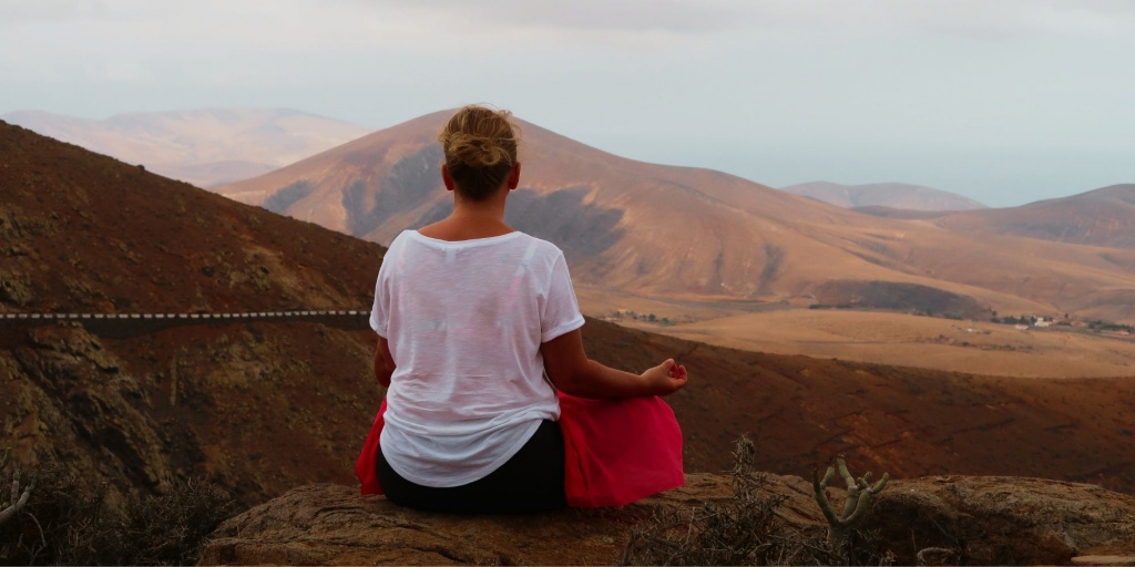 Daniela Brandl sitzt meditierend im Schneidersitz auf einem Felsen und blickt auf eine ruhige, weitläufige Berglandschaft, die Entspannung und innere Ruhe symbolisiert.