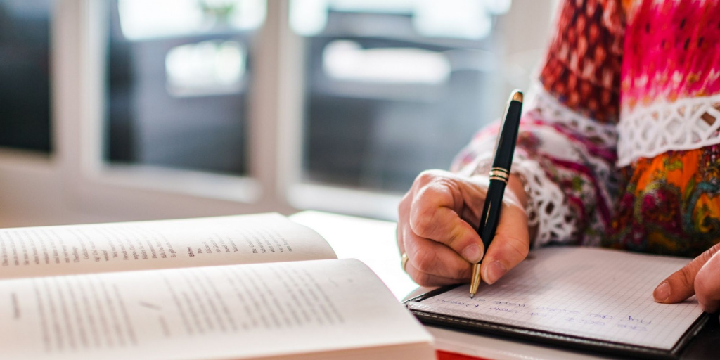 Nahaufnahme einer Hand, die mit einem Stift in ein Notizheft schreibt; daneben liegt ein aufgeschlagenes Buch, das Journaling und Selbstreflexion symbolisiert.