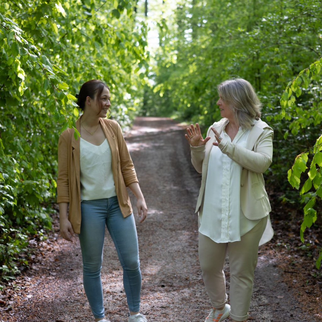 Daniela Brandl geht mit einer Coaching Klientin einen Waldweg entlang. Beide unterhalten sich angeregt. 