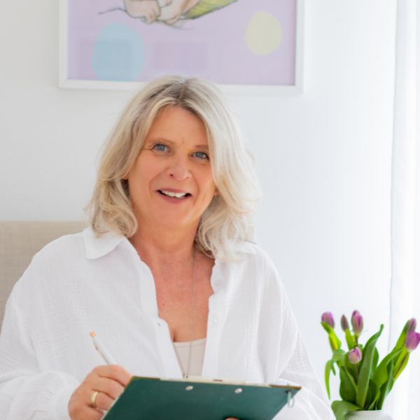 Daniela Brandl schaut lächelnd in die Kamera. In ihrer Hand hält sie einen Stift. Rechts im Bild sind lilafarbene Tulpen in einer Vase zu sehen.