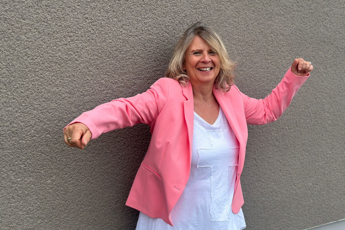 Daniela Brandl steht gut gelaunt vor einer erdfarbenen Wand, ihre Arme schwingen