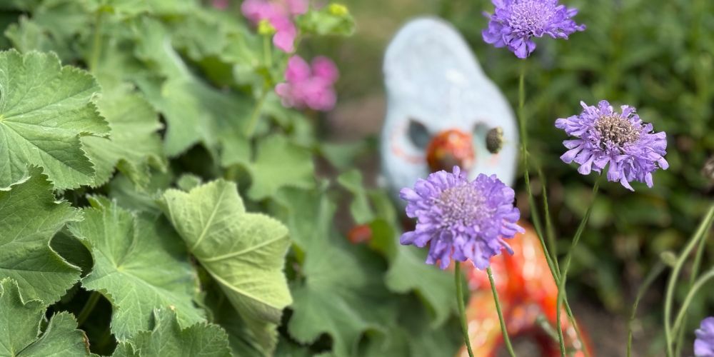 Eine bunte Tonfigur sitzt hinter violetten Blüten in einem grünen Beet