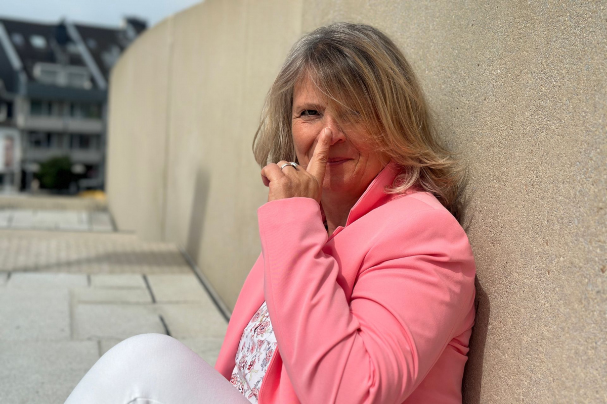 Daniela Brandl sitzt vor einer naturfarbenen Mauer, schaut lächelnd in die Kamera und hält ihren Zeigefinger an ihre Nasenspitze.