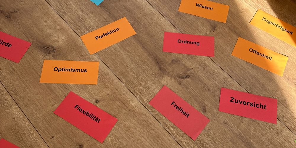 Rote und orangefarbene Moderationskarten, auf denen jeweils ein Wert steht, liegen auf einem Holzboden. Zu sehen sind: Perfektion, Freiheit, Zuversicht. Ordnung, Offenheit, Optimismus, Wissen, Würde